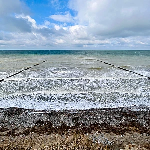 Im Vordergrund das Hohe Ufer, Richtung Horizont das stürmische Meer. Im oberen Drittel ein wolkenverhangener blauer Himmel.
