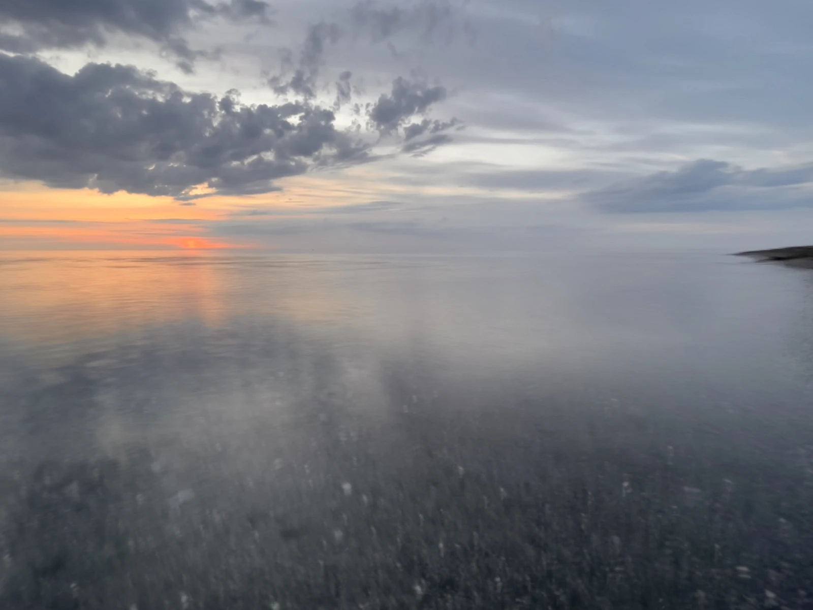 Sonnenuntergang. Das Bild ist gedrittelt. Zwei Drittel nimmt das durch die Langzeitbelichtung verschwommene Meer ein, ein Drittel die leicht orangene Sonne und bläulich graue Wolken am Himmel.