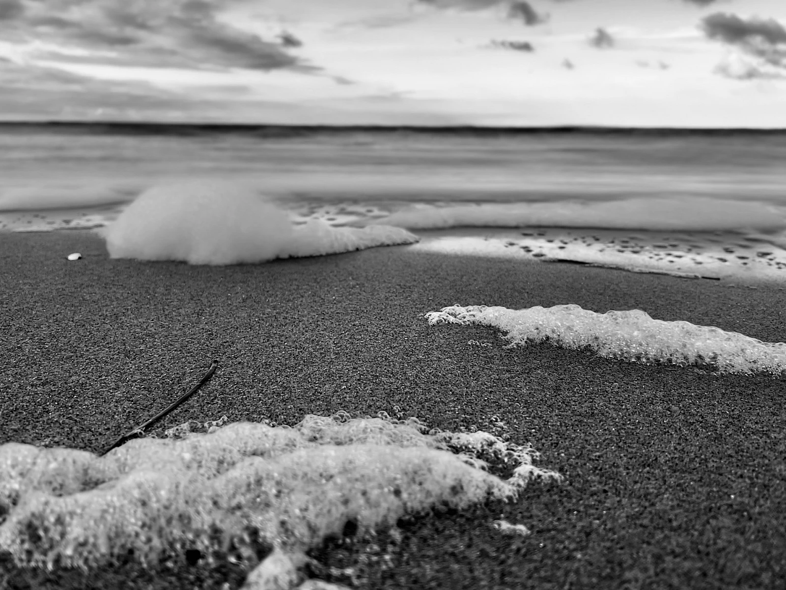Schwarzweiß-Aufnahme der Ostsee. Im Vordergrund sind Häufchen von Meerschaum auf dem Strand zu sehen.
