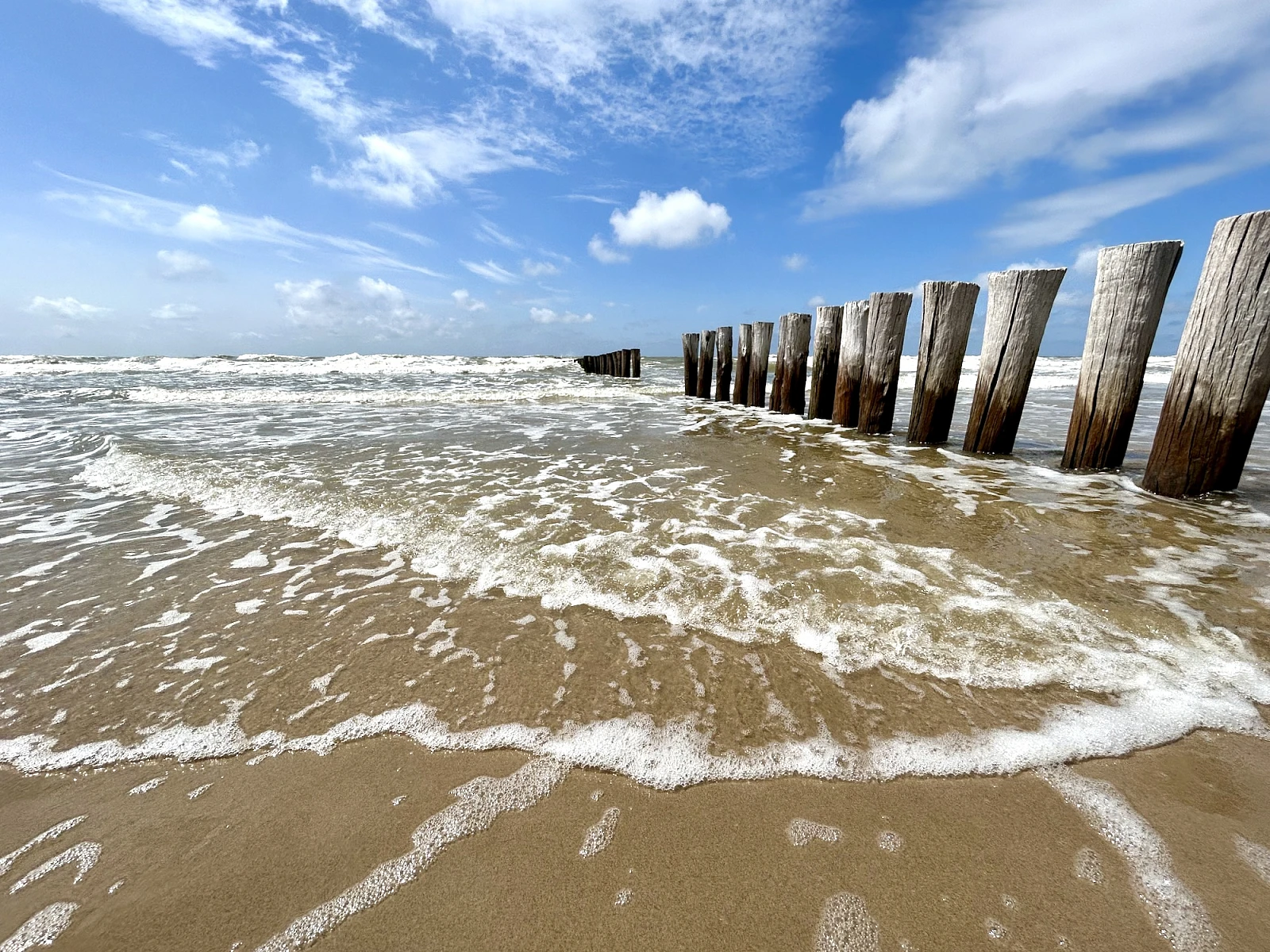 Rechts zieht sich vom Horizon zum Bildrand eine Reihe Holzbuhnen. Das Bild ist horizontal geteilt in ein Drittel Himmel, zwei Drittel Meer mit leichter auslaufender Brandung.
