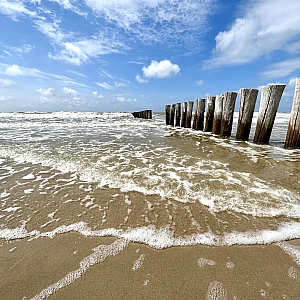 Rechts zieht sich vom Horizon zum Bildrand eine Reihe Holzbuhnen. Das Bild ist horizontal geteilt in ein Drittel Himmel, zwei Drittel Meer mit leichter auslaufender Brandung.