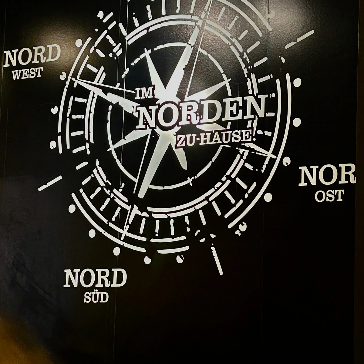 Foto eines Wandbildes. Auf schwarzem Grund ist ein Kompass zu sehen, dessen Richtungen Nord Nord, Nord Ost, Nord Süd und Nord West sind. Im Zentrum steht: Im Norden zu Hause