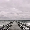 Im unteren Drittel des Bildes ragt eine Holzbrücke in das wilde grüne Meer. Oberhalb des Horizonts dichte Wolken. Alles ist grau.