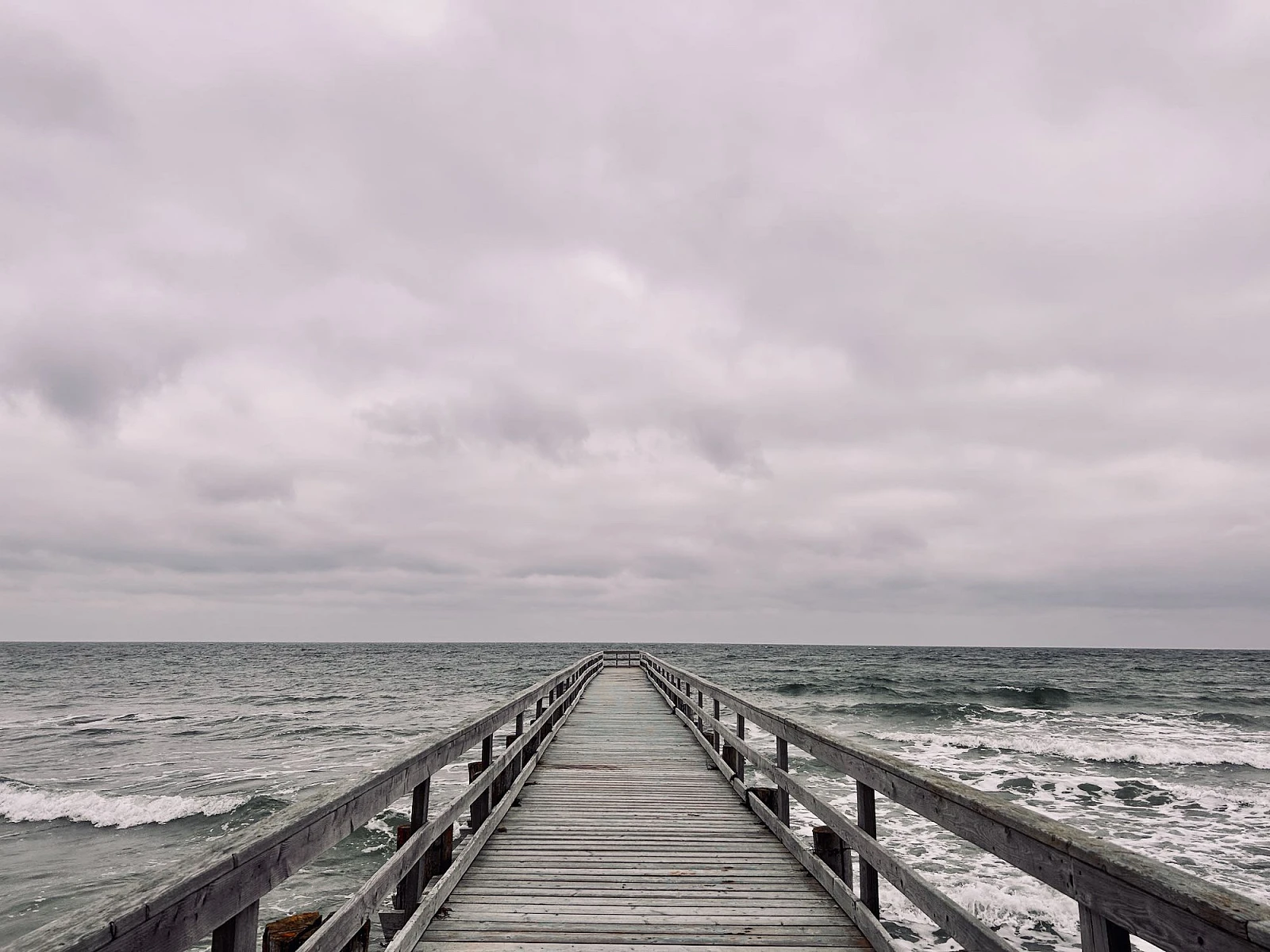 Im unteren Drittel des Bildes ragt eine Holzbrücke in das wilde grüne Meer. Oberhalb des Horizonts dichte Wolken. Alles ist grau.