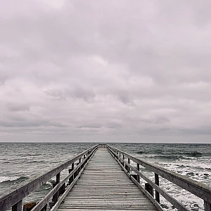 Im unteren Drittel des Bildes ragt eine Holzbrücke in das wilde grüne Meer. Oberhalb des Horizonts dichte Wolken. Alles ist grau.
