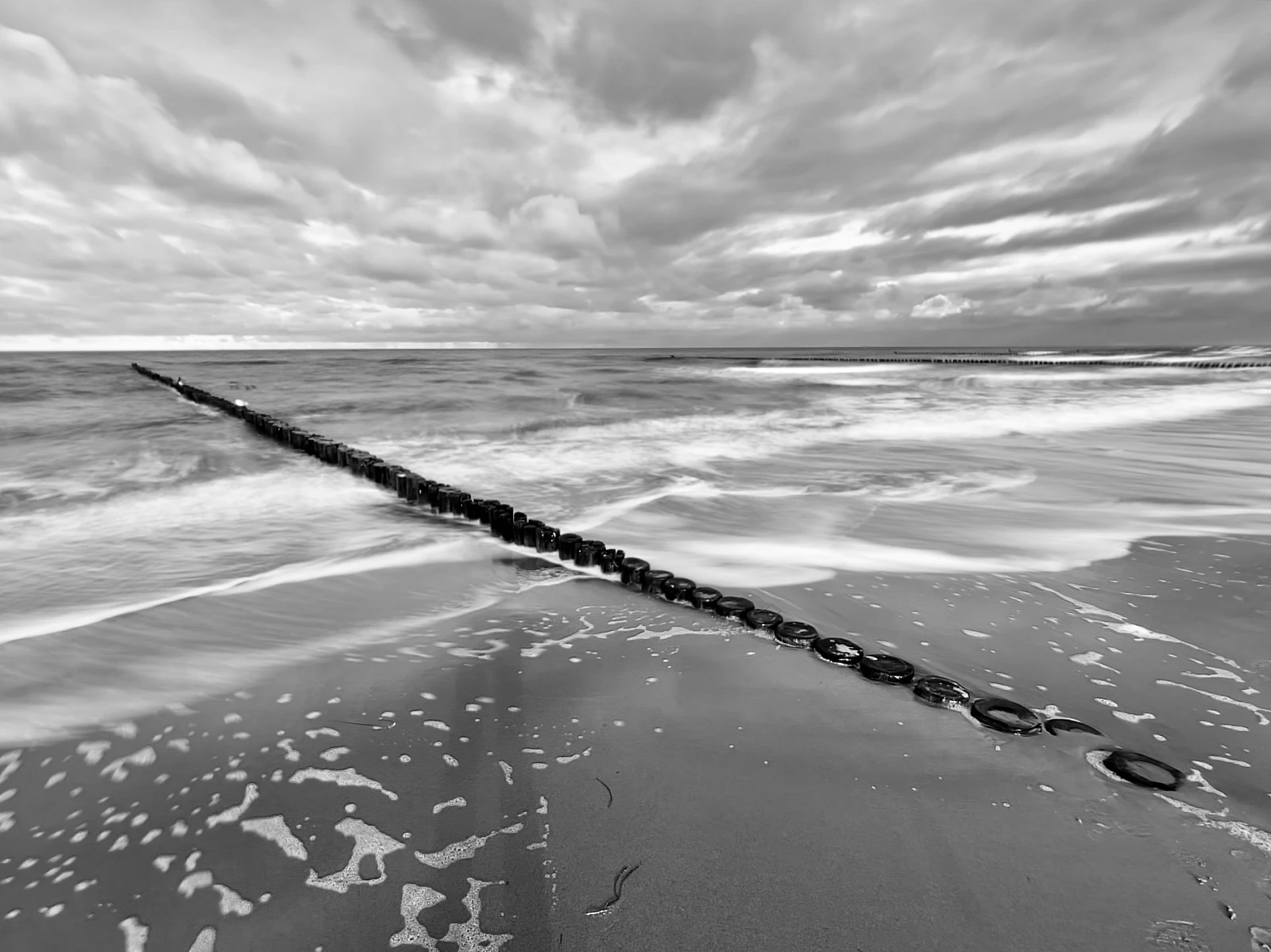 Schwarzweiß-Aufnahme der Ostsee. Eine Holzbuhne zieht sich diagonal von links oben nach rechts unten durchs Bild.