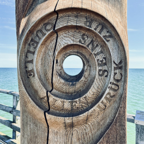 Blick durch eine Holzskulptur auf das Meer. Auf der Skulptur ist geschnitzt: Zurück zur Quelle allen Seins
