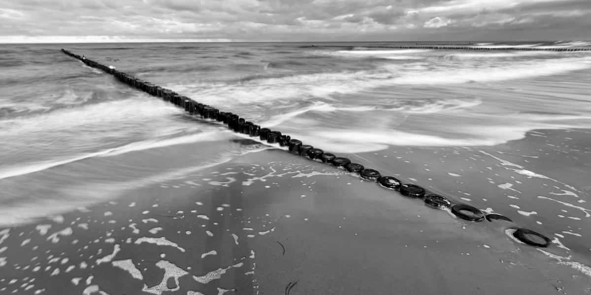 Schwarzweiß-Aufnahme der Ostsee. Eine Holzbuhne zieht sich diagonal von links oben nach rechts unten durchs Bild.