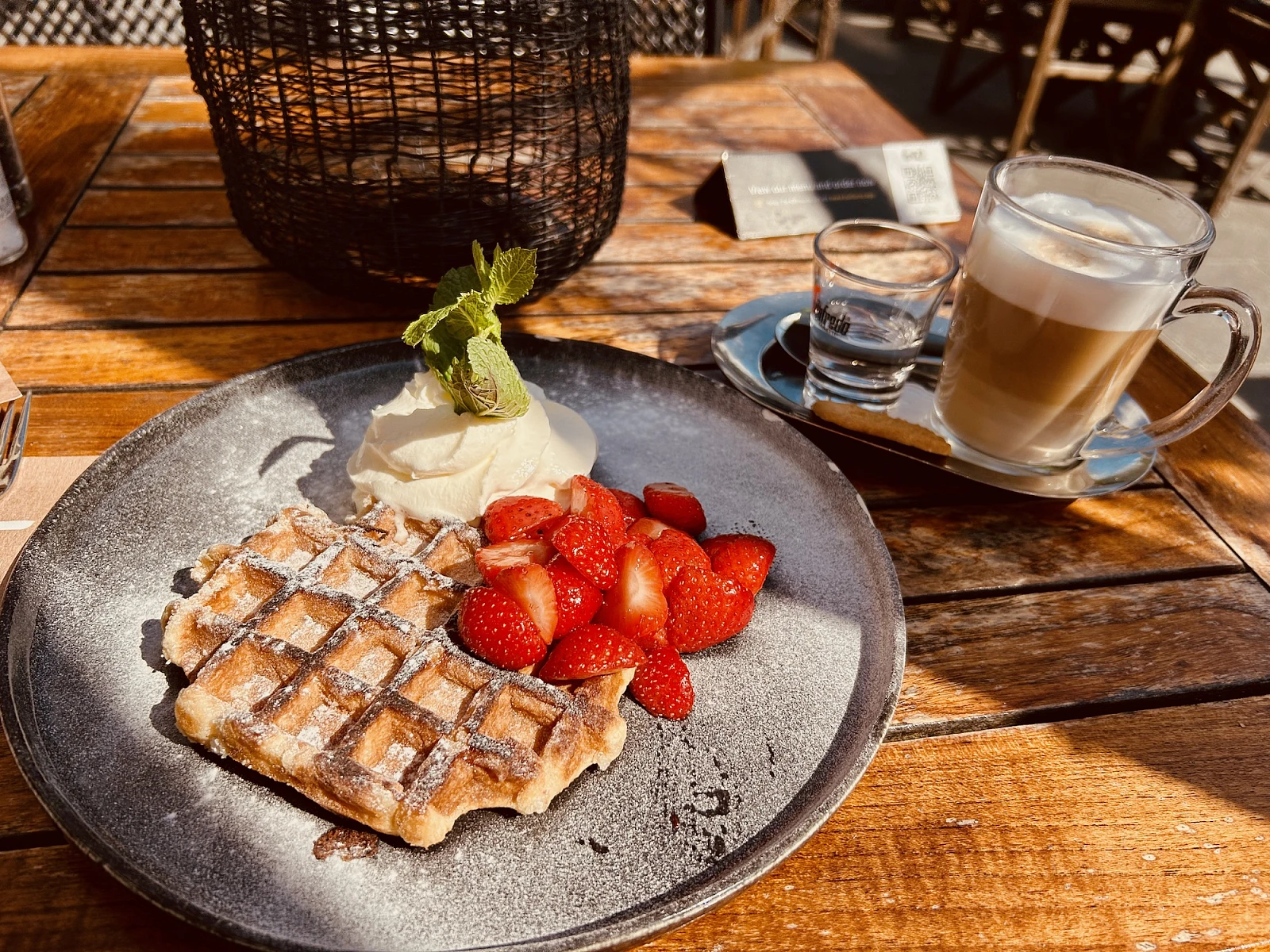 Auf einem Holztisch in warmen Sonnenlicht stehen ein Teller und eine Kaffeetasse mit kleinem Wasserglas. Auf dem Teller eine Belgische Waffel mit Erdbeeren und Schlagsahne.