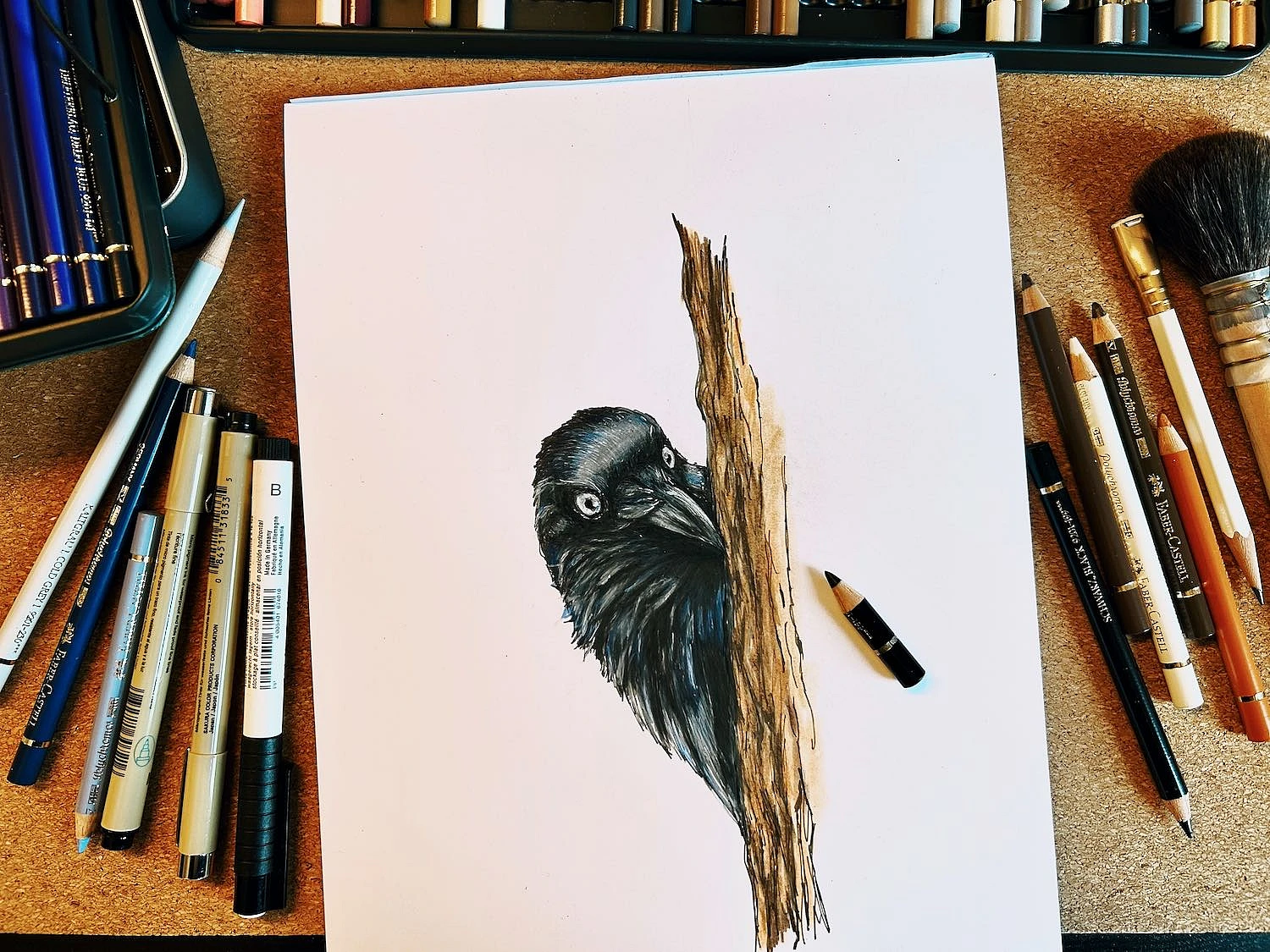 Auf einer Schreibtischunterlage aus Kork liegt mittig die Zeichnung einer Dohle. Rings herum sind Utensilien verteilt: Polychromos, Fineliner, Bleistifte und ein Pinsel.