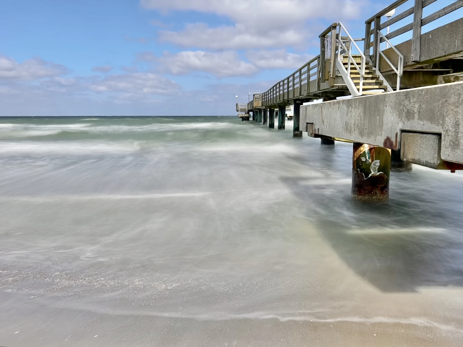 Die Seebrücke von Rerik ist am rechten Rand angeschnitten. Sie ist umgeben vom durch die Langzeitbelichtung weich gewaschen Meer. Der Himmel ist blau mit kleinen weißen Wolken.