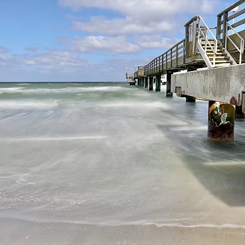Die Seebrücke von Rerik ist am rechten Rand angeschnitten. Sie ist umgeben vom durch die Langzeitbelichtung weich gewaschen Meer. Der Himmel ist blau mit kleinen weißen Wolken.