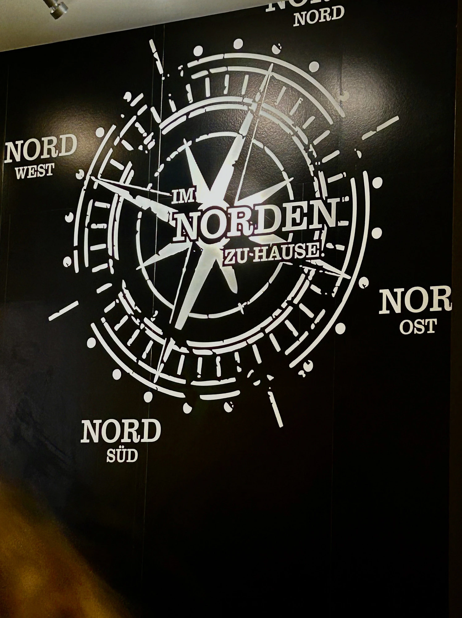Foto eines Wandbildes. Auf schwarzem Grund ist ein Kompass zu sehen, dessen Richtungen Nord Nord, Nord Ost, Nord Süd und Nord West sind. Im Zentrum steht: Im Norden zu Hause