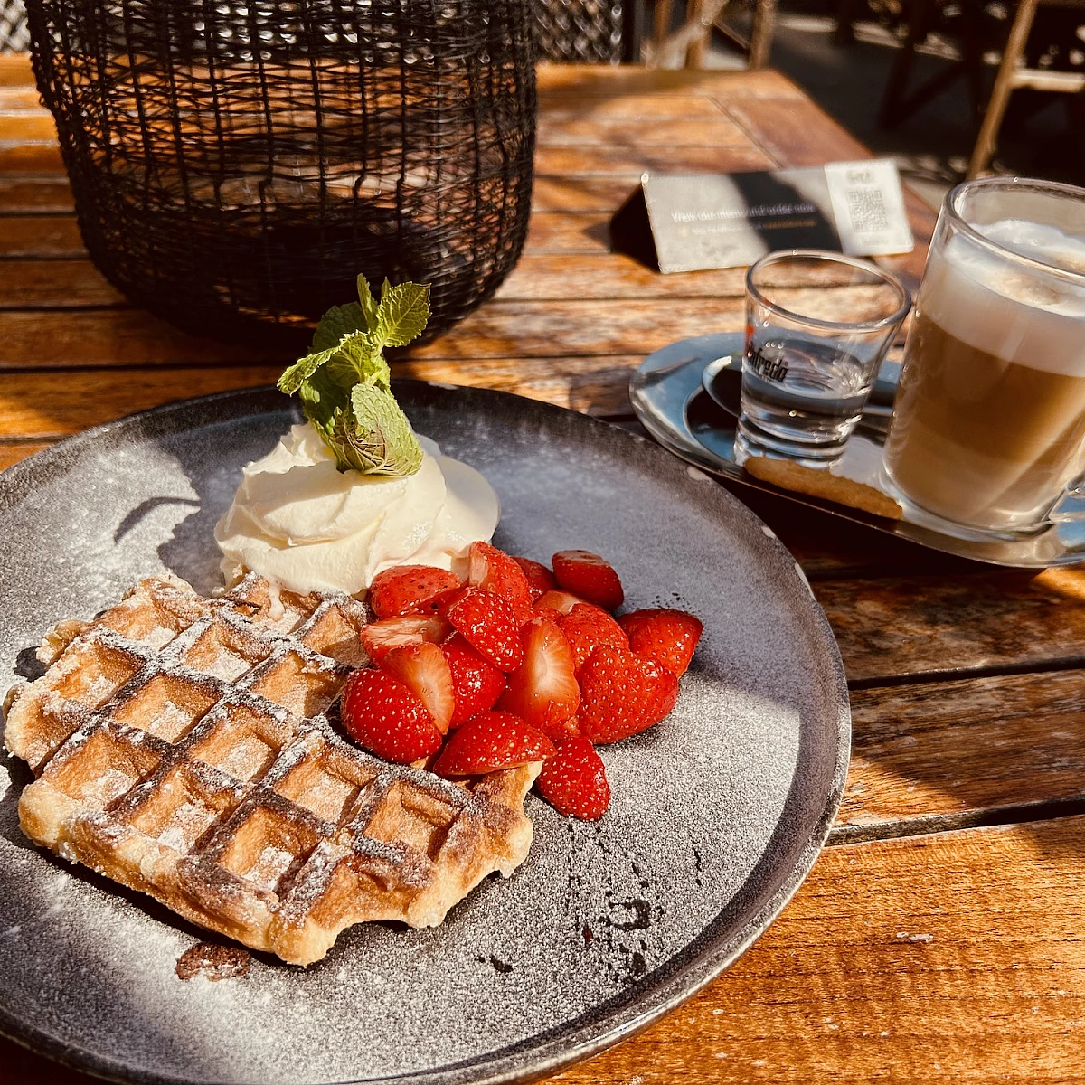 Auf einem Holztisch in warmen Sonnenlicht stehen ein Teller und eine Kaffeetasse mit kleinem Wasserglas. Auf dem Teller eine Belgische Waffel mit Erdbeeren und Schlagsahne.