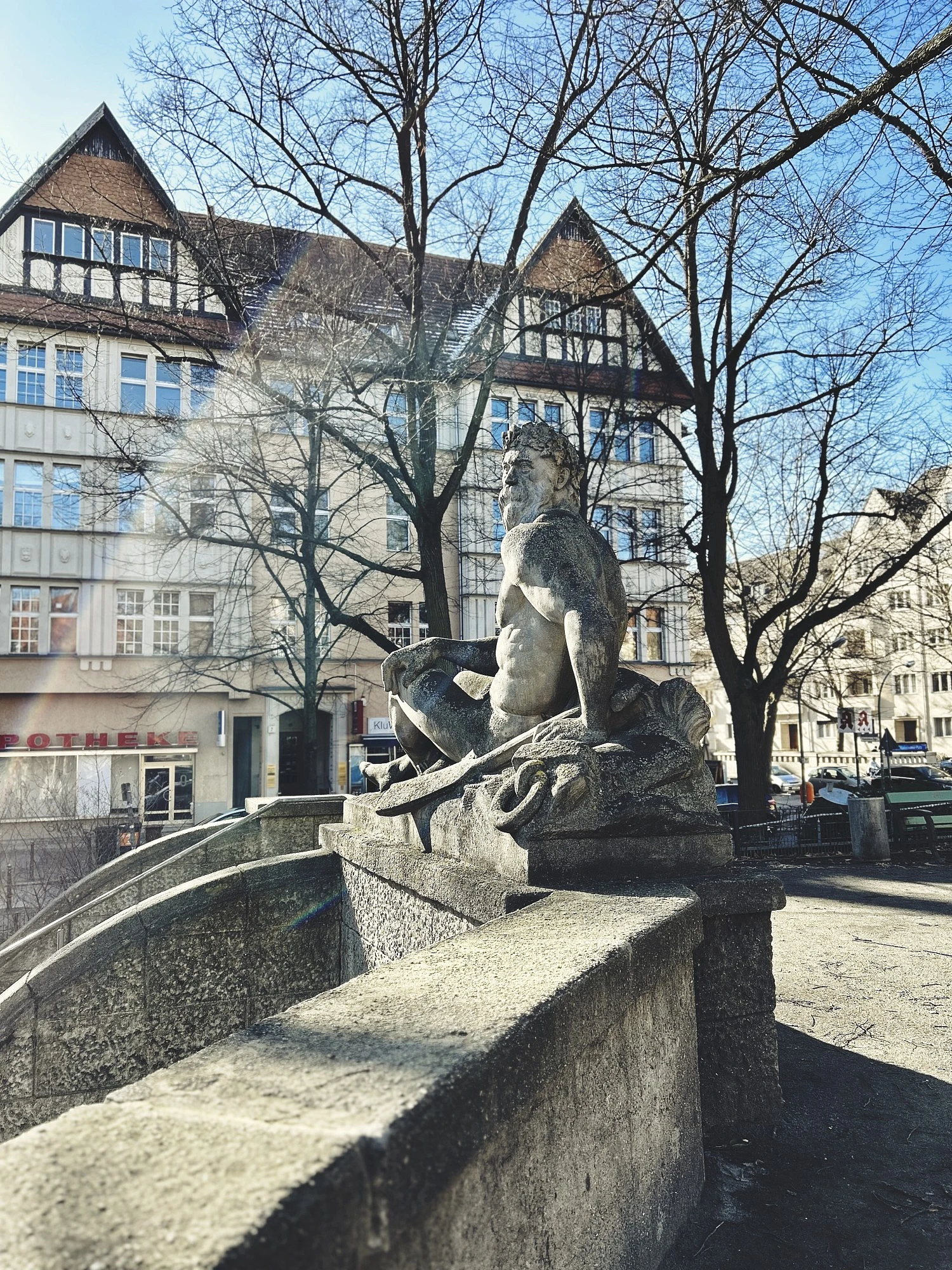 Die Statue eines sitzenden Mannes im Vordergrund, im Hintergrun ein Haus mit zwei spitzen Giebeln.