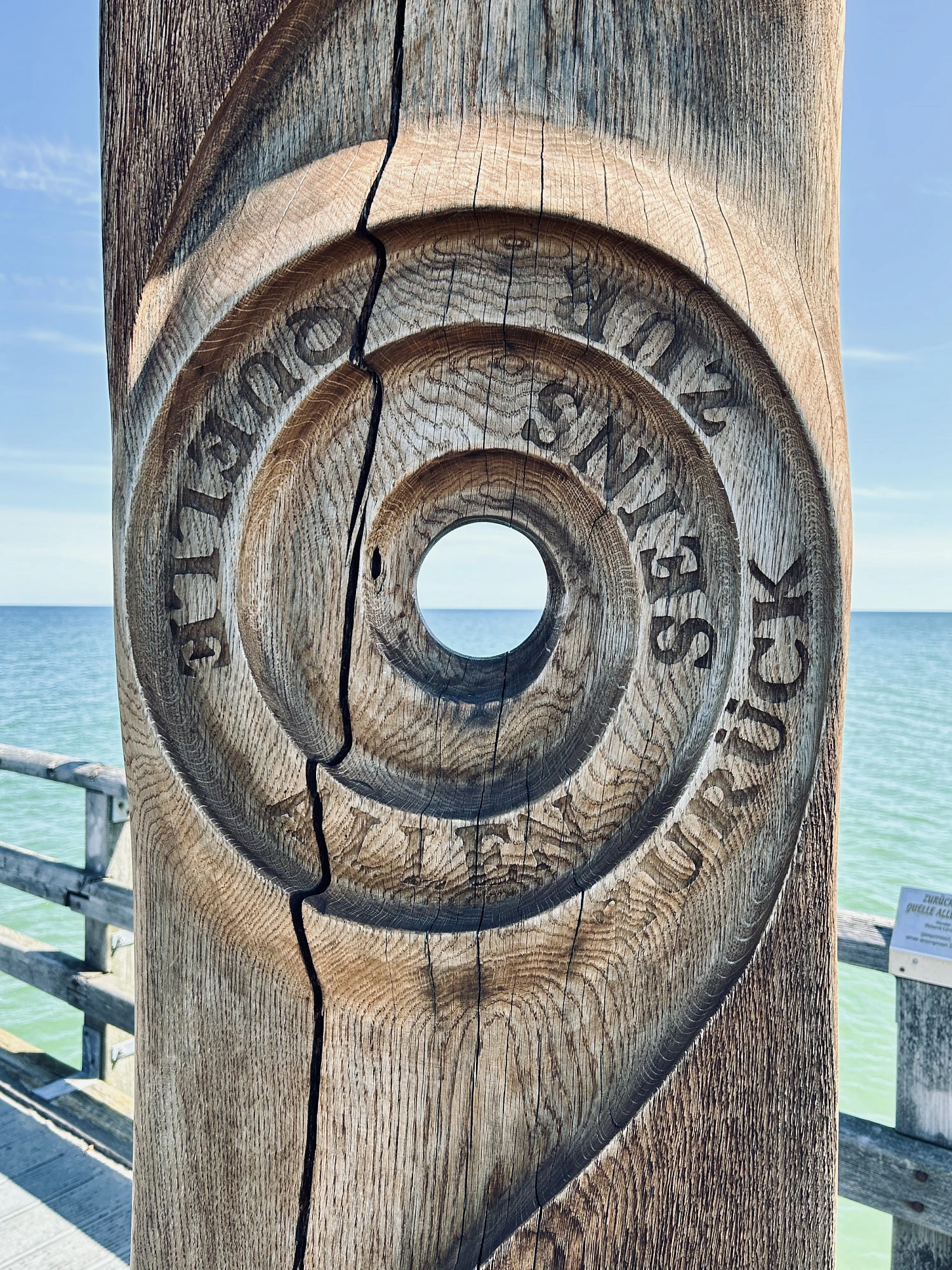 Blick durch eine Holzskulptur auf das Meer. Auf der Skulptur ist geschnitzt: Zurück zur Quelle allen Seins