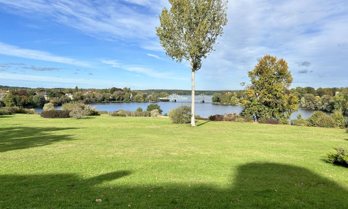 Das Bild zeigt eine malerische Landschaft mit einer weitläufigen, grünen Wiese im Vordergrund. Auf der Wiese stehen zwei Bäume, einer mit grünen Blättern und der andere mit herbstlich gefärbten Blättern. Im Hintergrund sieht man einen ruhigen See, umgeben von Bäumen und Sträuchern. Jenseits des Sees sind Häuser und eine Brücke zu erkennen. Der Himmel ist blau mit einigen weißen Wolken. Es sieht nach einem sonnigen Tag aus.