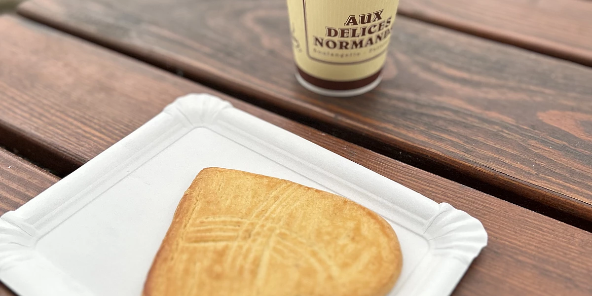 Foto. Auf einem Holztisch im Freien steht ein Pappbecher mit Kaffee. Darunter liegt auf einem Pappteller ein herzförmiger großer Cookie.