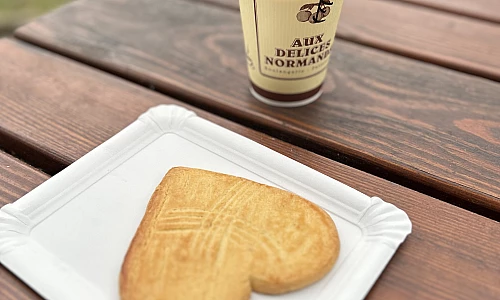 Foto. Auf einem Holztisch im Freien steht ein Pappbecher mit Kaffee. Darunter liegt auf einem Pappteller ein herzförmiger großer Cookie.
