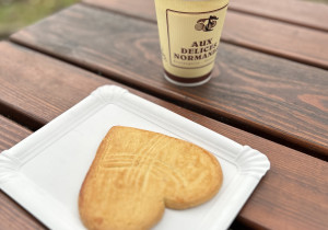 Foto. Auf einem Holztisch im Freien steht ein Pappbecher mit Kaffee. Darunter liegt auf einem Pappteller ein herzförmiger großer Cookie.