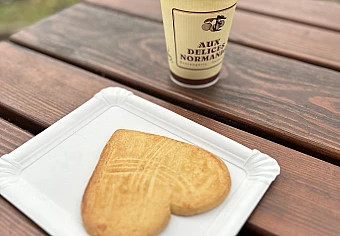 Foto. Auf einem Holztisch im Freien steht ein Pappbecher mit Kaffee. Darunter liegt auf einem Pappteller ein herzförmiger großer Cookie.