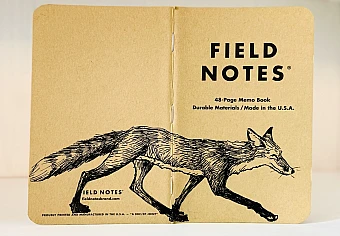 Auf dem aufgeklappten Rücken eines Field Notes Heftes ist eine schwarze Tintezeichnung eines Fuchses zu sehen. Der sehr schlanke Fuchs läuft von links nach rechts.