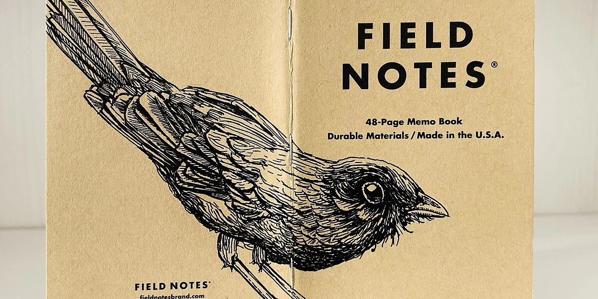 Foto eines stehenden und aufgeklappten Field Notes Heftes. Über den Rücken erstreckt sich die schwarze Tintezeichnung einer Amsel, die nach rechts schaut.