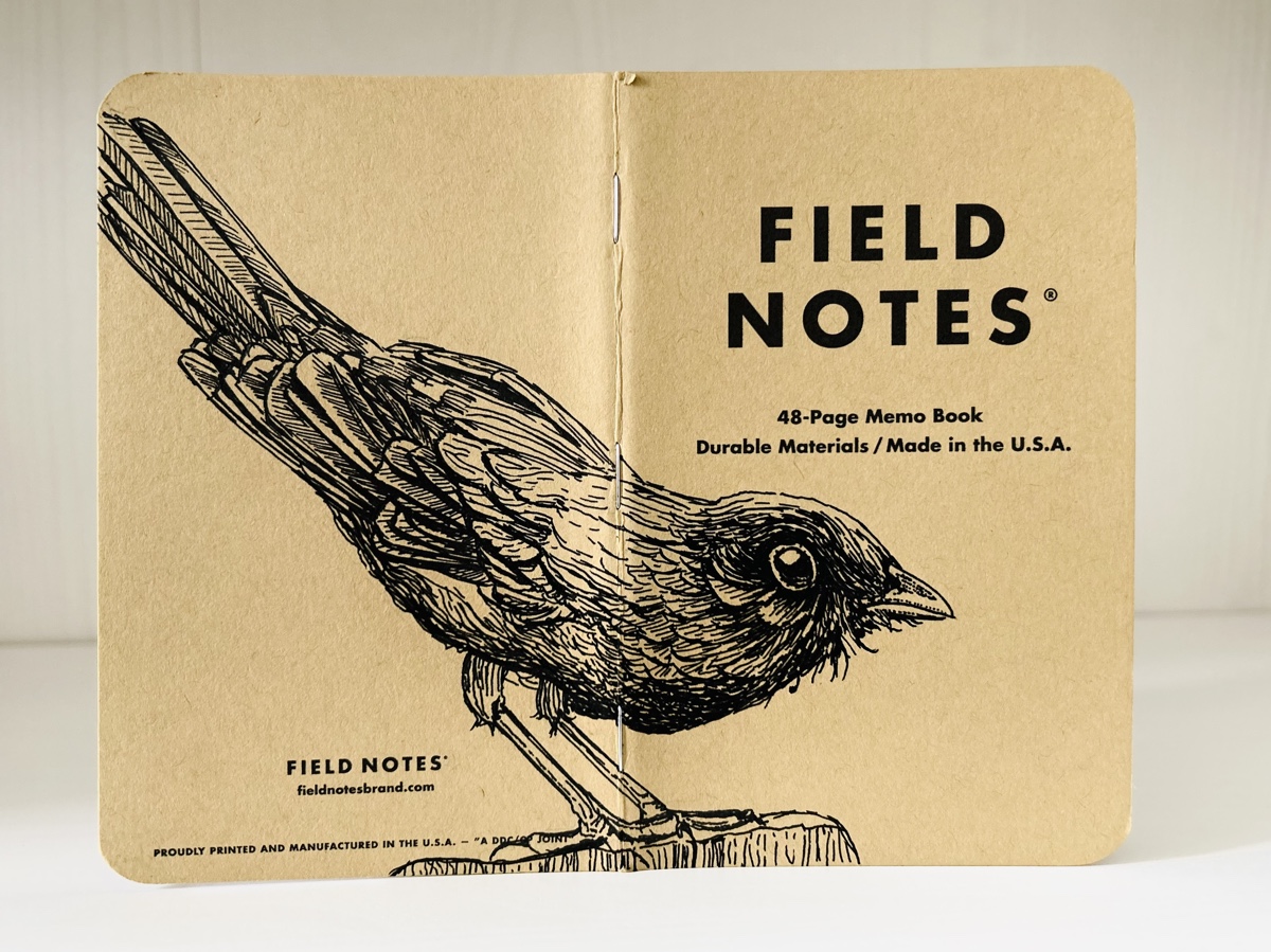Foto eines stehenden und aufgeklappten Field Notes Heftes. Über den Rücken erstreckt sich die schwarze Tintezeichnung einer Amsel, die nach rechts schaut.