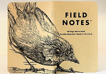Foto eines stehenden und aufgeklappten Field Notes Heftes. Über den Rücken erstreckt sich die schwarze Tintezeichnung eines Rotkehlchens, das geduckt nach rechts schaut.