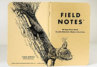Foto eines stehenden und aufgeklappten Field Notes Heftes. Über den Rücken erstreckt sich die schwarze Tintezeichnung eines kleinen Eichhörnchens, das an einem Baum abwärts klettert und den Kopf nach vorn streckt.