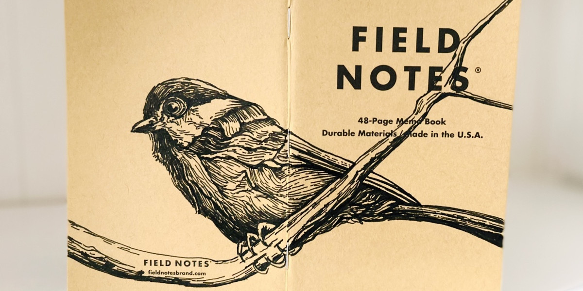 Foto eines stehenden und aufgeklappten Field Notes Heftes. Über den Rücken erstreckt sich die schwarze Tintezeichnung einer kleinen Meise, die auf einem Ast sitzt.