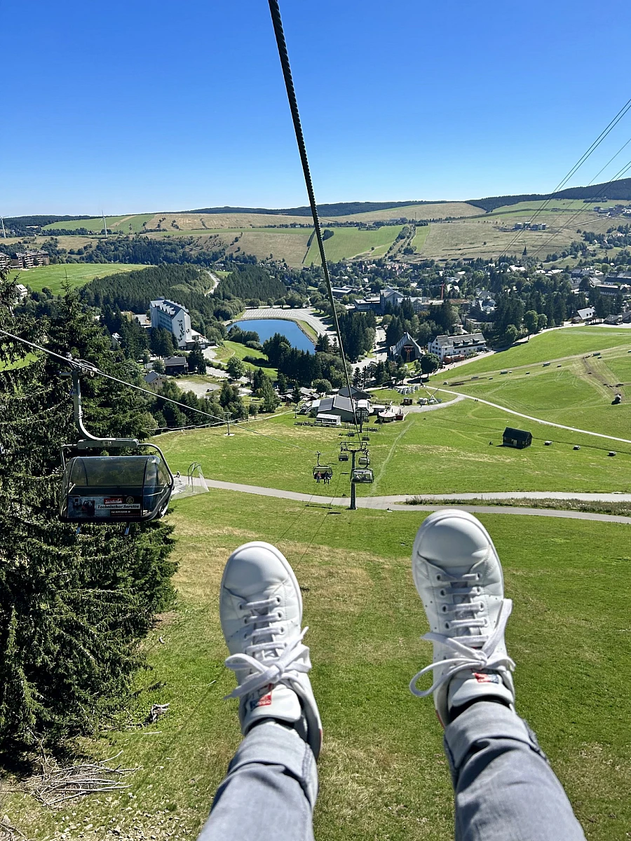 Aus einer Seilbahngondel nach unten fotografiert: Jeans und Sneaker über einer Wiese. Im Hintergrund und Tal Häuser und Felder.