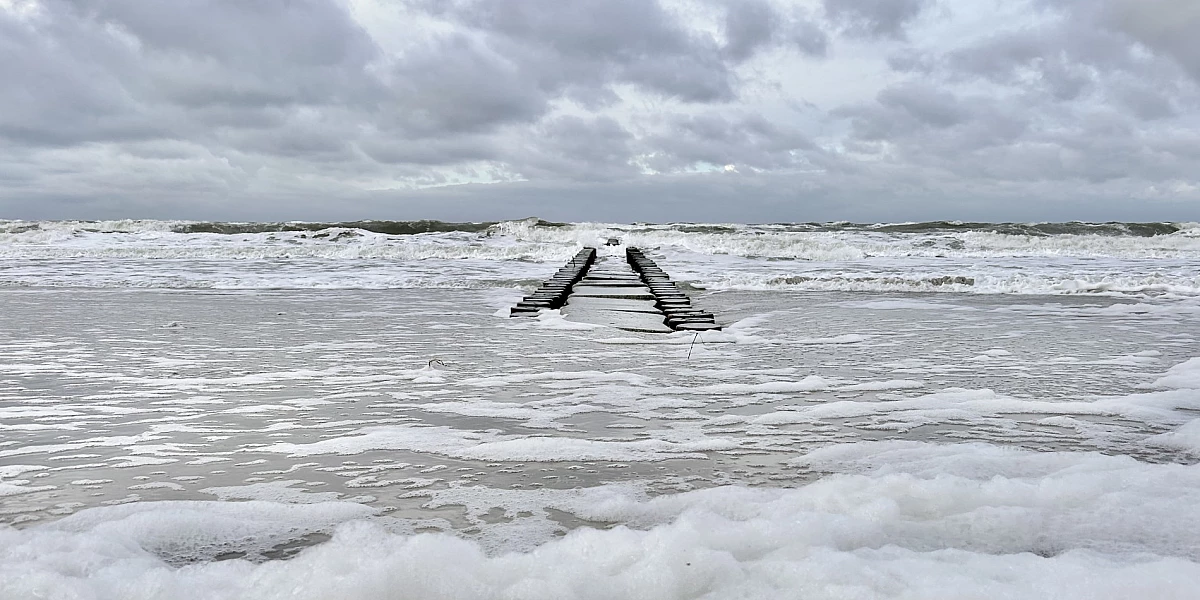 Foto der Ostsee. Eine Holzbuhne versinkt in Wellen, am Stand liegt Schaum.