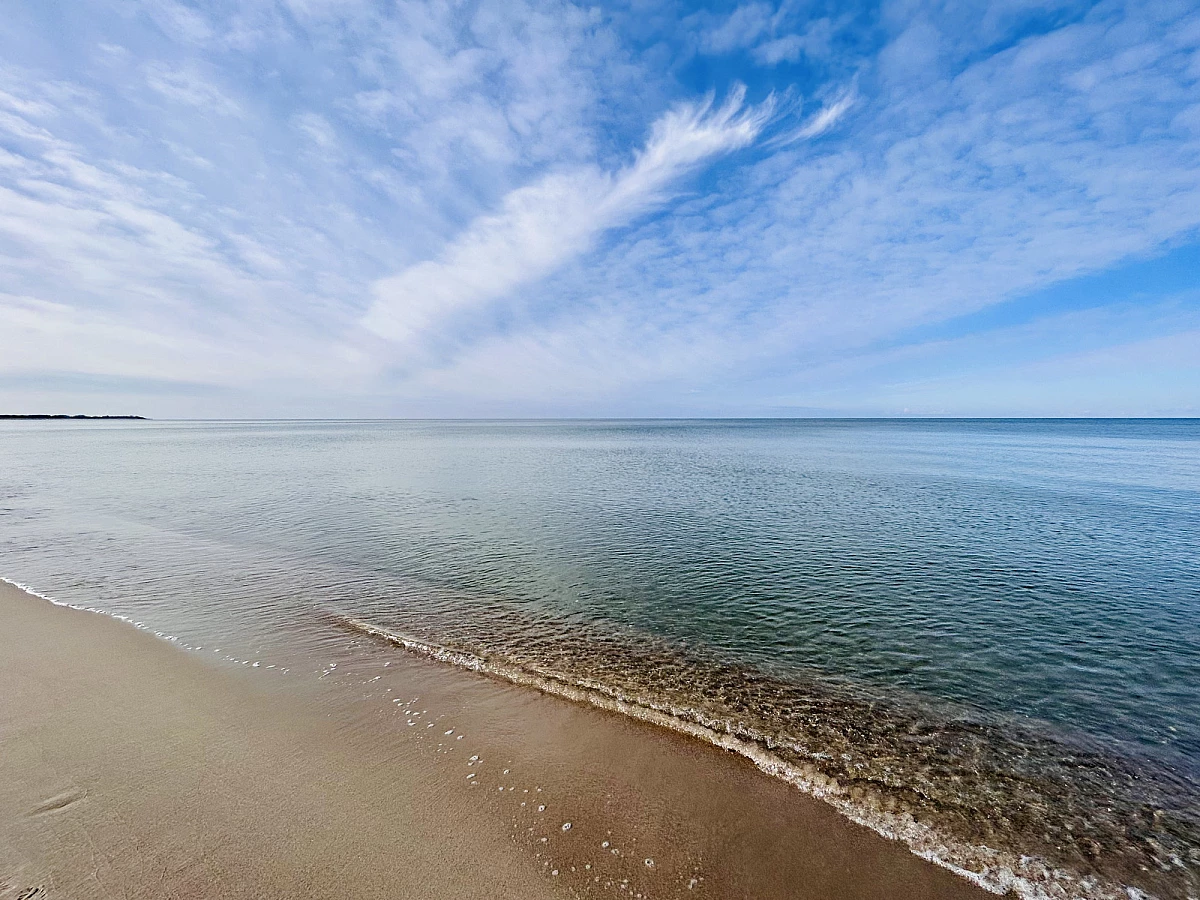 Das Bild ist zweigeteilt. Über dem Horizon der blaue Himmel mit weißen Wolkenstreifen. Im unteren Teil des Bildes das stille blaugrüne Meer und ganz links etwas Sandstrand.