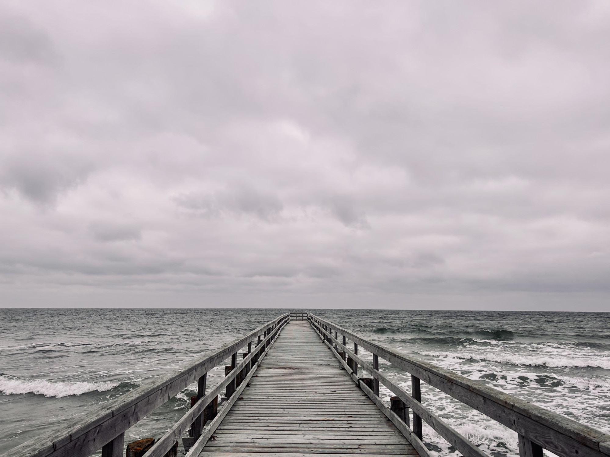 Ein grauer wolkenverhangener Himmel über dem unruhigen blaugrünen Meer.Im Vordergrund bis zum Horizont eine graue Holzbrücke.