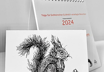 Der Kalender "Ommmmmmmmmmmm - Yoga für Eichhörnchen & ähnlich veranlagte Menschen - Postkartenkalender 2024"  steht aufgeklappt auf einer weißen Fläche. Im Vordergrund eine abgetrennte Postkarte.