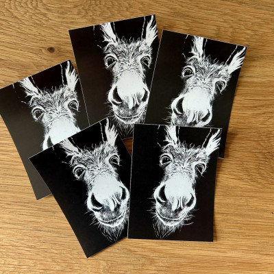 Foto von fünf Aufklebern, die auf einem Holztisch liegen. Der Aufkleber selbst zeigt die weiße Zeichnung eines freundlichen dreinschauenden Esels auf schwarzem Grund.