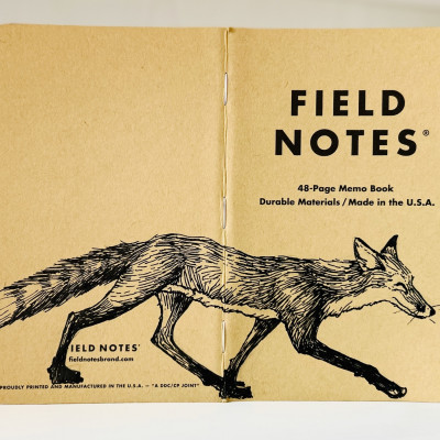Auf dem aufgeklappten Rücken eines Field Notes Heftes ist eine schwarze Tintezeichnung eines Fuchses zu sehen. Der sehr schlanke Fuchs läuft von links nach rechts.