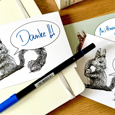 Auf einem Holztisch liegt ein Notizbuch und ein Stift. Darüber liegen Postkarten mit dem Motiv eines Eichhörnchens mit einer Sprechblase. In den Sprechblasen sind handschriftlich die Schriftzüge "Danke!", "Mittwoch um 9.00?" und "Hi,".