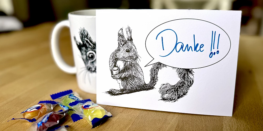 Eine Postkarte mit dem Motiv eines Eichhörnchens mit Sprechblase steht aufrecht gegen eine Tasse gelehnt auf einem Holztisch. Davor ein paar Bonbons. Auf der Karte steht handschriftlich 