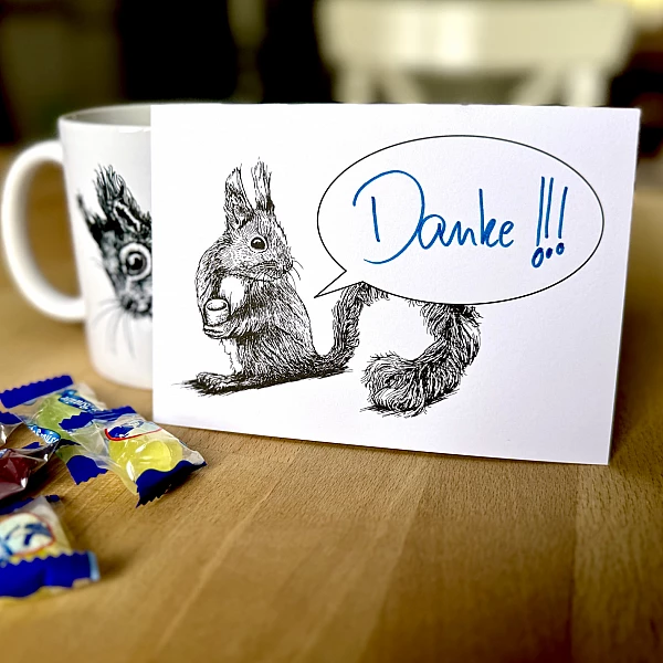 Eine Postkarte mit dem Motiv eines Eichhörnchens mit Sprechblase steht aufrecht gegen eine Tasse gelehnt auf einem Holztisch. Davor ein paar Bonbons. Auf der Karte steht handschriftlich "Danke!!!".