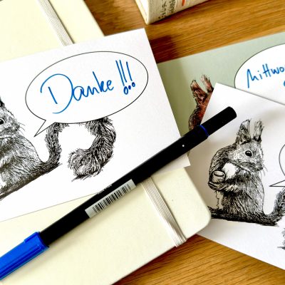 Auf einem Holztisch liegt ein Notizbuch und ein Stift. Darüber liegen Postkarten mit dem Motiv eines Eichhörnchens mit einer Sprechblase. In den Sprechblasen sind handschriftlich die Schriftzüge "Danke!", "Mittwoch um 9.00?" und "Hi,".