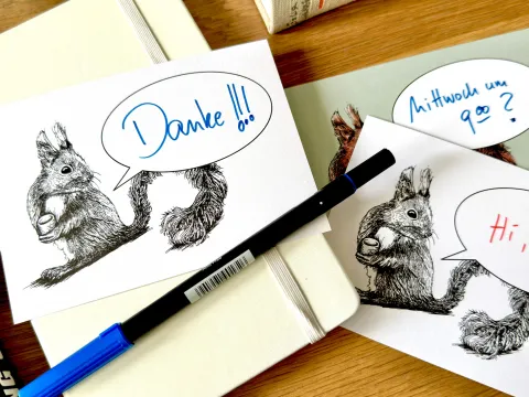 Auf einem Holztisch liegt ein Notizbuch und ein Stift. Darüber liegen Postkarten mit dem Motiv eines Eichhörnchens mit einer Sprechblase. In den Sprechblasen sind handschriftlich die Schriftzüge "Danke!", "Mittwoch um 9.00?" und "Hi,".