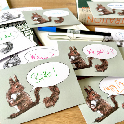 Verschiedene Postkarten mit mit dem Motiv eines Eichhörnchen und einer Sprechblase liegen auf einen Holztisch. In den Sprechblasen finden sich handschriftlich Schriftzüge wie "Bitte!", "Wie geht's?", "Wann?", "Happy Monday"