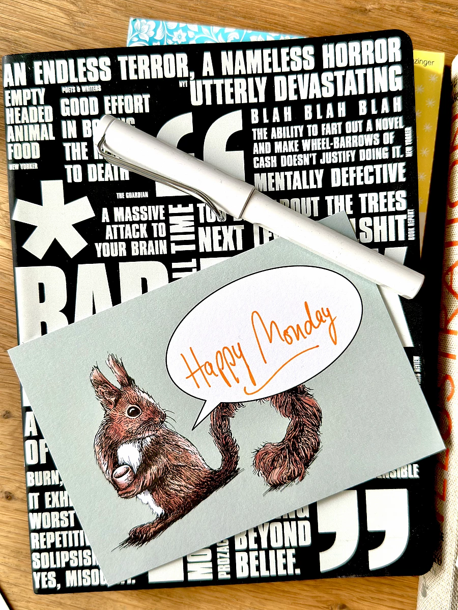 Eine Postkarte mit dem Motiv eines Eichhörnchens und einer Sprechblase und dem handschriftlichen Schriftzug "Happy Monday" liegt zusammen mit einem Füllhalter auf einem Notizbuch auf einem Holztisch.