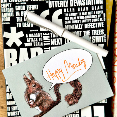 Eine Postkarte mit dem Motiv eines Eichhörnchens und einer Sprechblase und dem handschriftlichen Schriftzug "Happy Monday" liegt zusammen mit einem Füllhalter auf einem Notizbuch auf einem Holztisch.