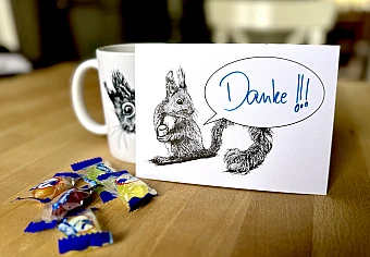 Eine Postkarte mit dem Motiv eines Eichhörnchens mit Sprechblase steht aufrecht gegen eine Tasse gelehnt auf einem Holztisch. Davor ein paar Bonbons. Auf der Karte steht handschriftlich "Danke!!!".