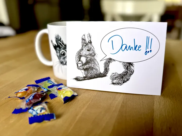 Eine Postkarte mit dem Motiv eines Eichhörnchens mit Sprechblase steht aufrecht gegen eine Tasse gelehnt auf einem Holztisch. Davor ein paar Bonbons. Auf der Karte steht handschriftlich "Danke!!!".