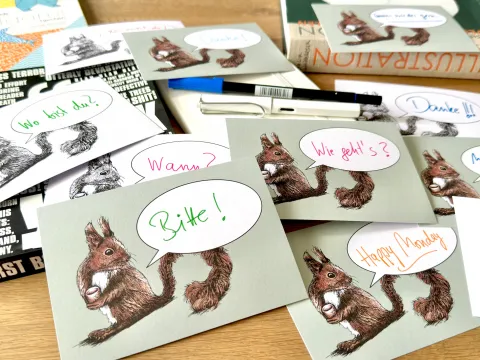 Verschiedene Postkarten mit mit dem Motiv eines Eichhörnchen und einer Sprechblase liegen auf einen Holztisch. In den Sprechblasen finden sich handschriftlich Schriftzüge wie "Bitte!", "Wie geht's?", "Wann?", "Happy Monday"