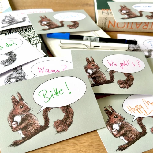 Verschiedene Postkarten mit mit dem Motiv eines Eichhörnchen und einer Sprechblase liegen auf einen Holztisch. In den Sprechblasen finden sich handschriftlich Schriftzüge wie 
